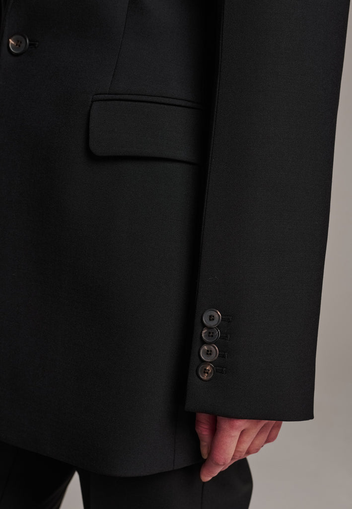 Experience flawless tailoring with this structured blazer, featuring sharp wide shoulders and a subtly cinched waist. Fully satin-lined for added luxury, it includes horn buttons and welt pockets with flaps, along with a back slit for ease of movement. Masterfully crafted in a versatile black hue, this blazer seamlessly complements a range of colors, embodying precision and elegance.