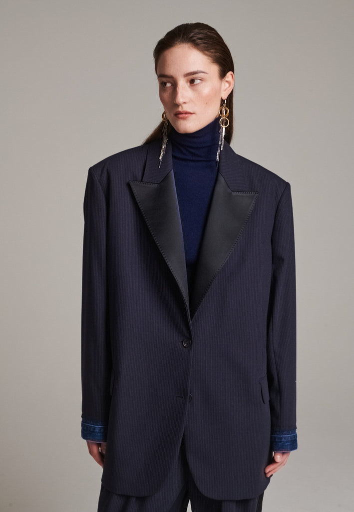 The Devotion Pinstripe Blazer Navy is a bold, reimagined take on classic tailoring. Designed with an oversized silhouette, it features a timeless navy pinstripe base contrasted by sleek black lapels in a subtly glossy fabric, adding a refined, modern twist. The cuffs break convention with a denim finish, introducing a casual edge to the otherwise structured piece. 