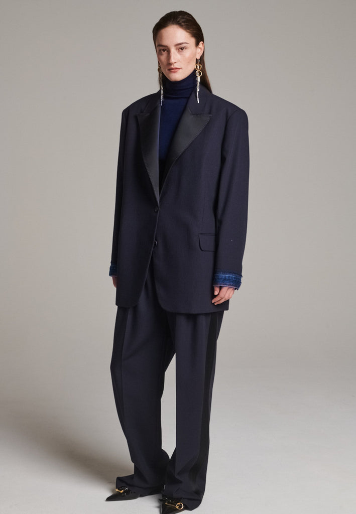 The Devotion Pinstripe Blazer Navy is a bold, reimagined take on classic tailoring. Designed with an oversized silhouette, it features a timeless navy pinstripe base contrasted by sleek black lapels in a subtly glossy fabric, adding a refined, modern twist. The cuffs break convention with a denim finish, introducing a casual edge to the otherwise structured piece. 