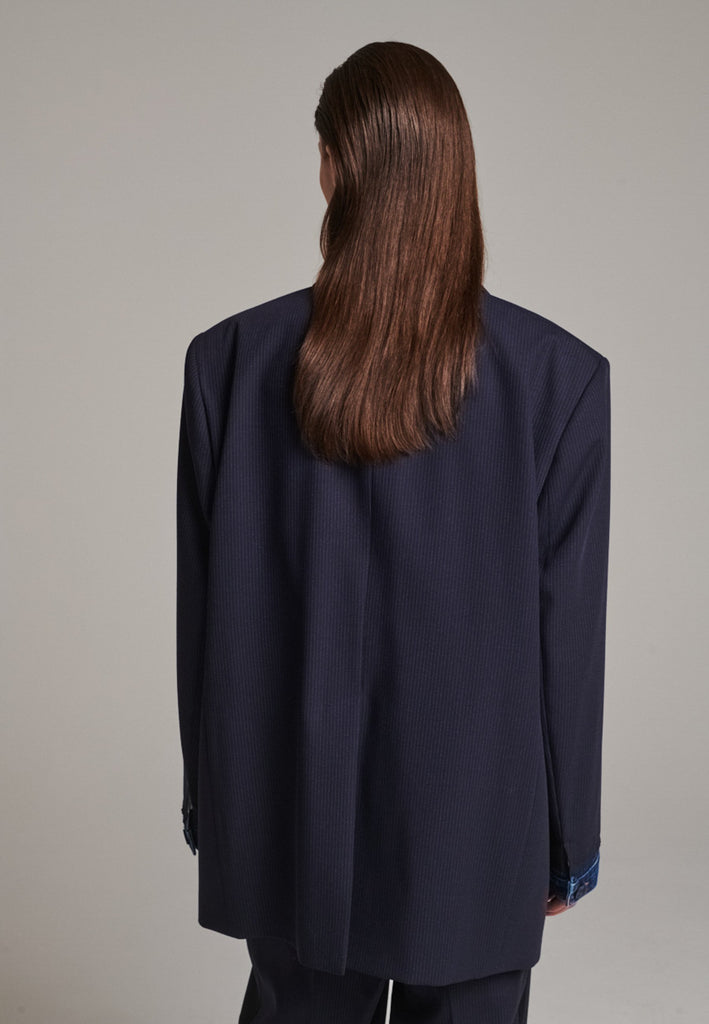 The Devotion Pinstripe Blazer Navy is a bold, reimagined take on classic tailoring. Designed with an oversized silhouette, it features a timeless navy pinstripe base contrasted by sleek black lapels in a subtly glossy fabric, adding a refined, modern twist. The cuffs break convention with a denim finish, introducing a casual edge to the otherwise structured piece. 