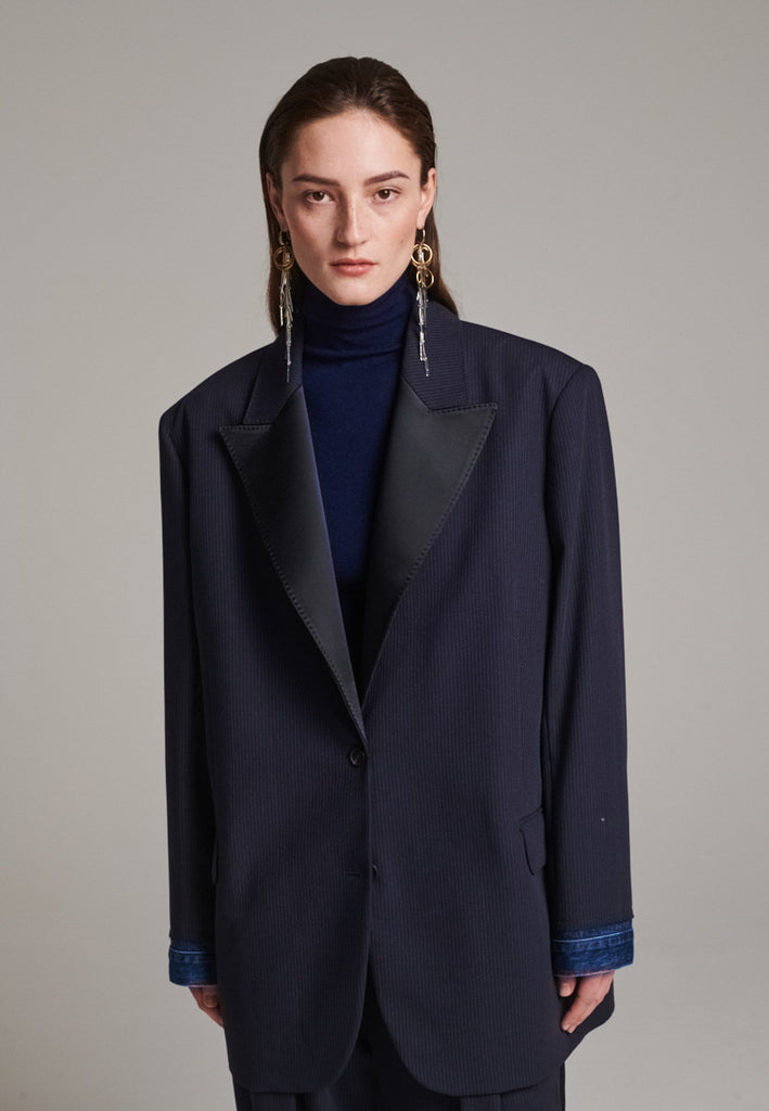 The Devotion Pinstripe Blazer Navy is a bold, reimagined take on classic tailoring. Designed with an oversized silhouette, it features a timeless navy pinstripe base contrasted by sleek black lapels in a subtly glossy fabric, adding a refined, modern twist. The cuffs break convention with a denim finish, introducing a casual edge to the otherwise structured piece. 