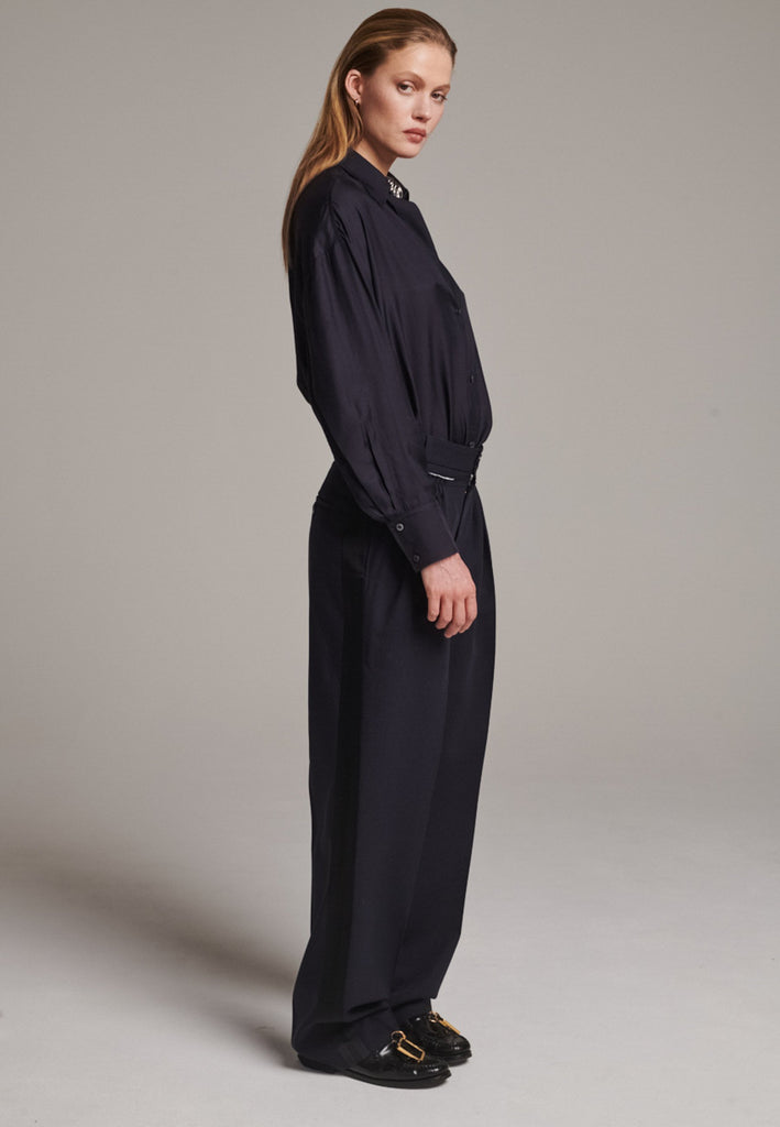 Straight-leg trousers with a classic cut, elevated by subtle yet striking details. The unfinished waistband adds a raw, intentional edge, contrasting with the otherwise clean silhouette. Glossy black fabric stripe runs down the side seams, bringing a touch of texture and definition to the navy pinstripe base. A refined everyday piece with a modern, deconstructed twist.