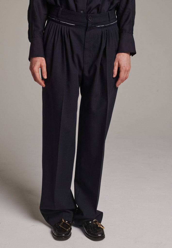 Straight-leg trousers with a classic cut, elevated by subtle yet striking details. The unfinished waistband adds a raw, intentional edge, contrasting with the otherwise clean silhouette. Glossy black fabric stripe runs down the side seams, bringing a touch of texture and definition to the navy pinstripe base. A refined everyday piece with a modern, deconstructed twist.