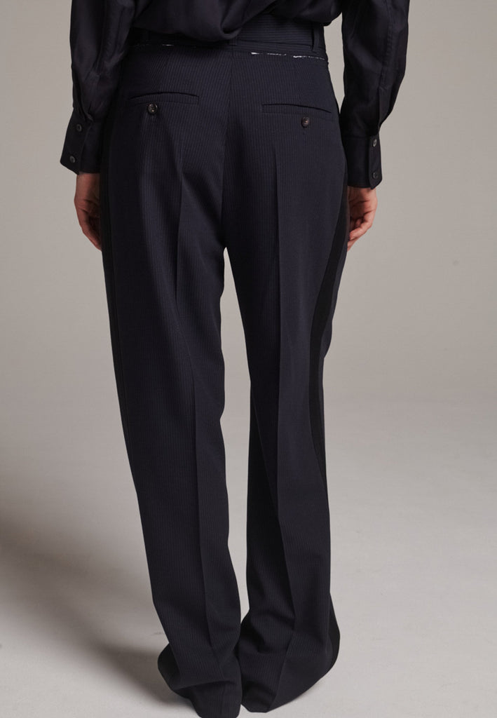 Straight-leg trousers with a classic cut, elevated by subtle yet striking details. The unfinished waistband adds a raw, intentional edge, contrasting with the otherwise clean silhouette. Glossy black fabric stripe runs down the side seams, bringing a touch of texture and definition to the navy pinstripe base. A refined everyday piece with a modern, deconstructed twist.