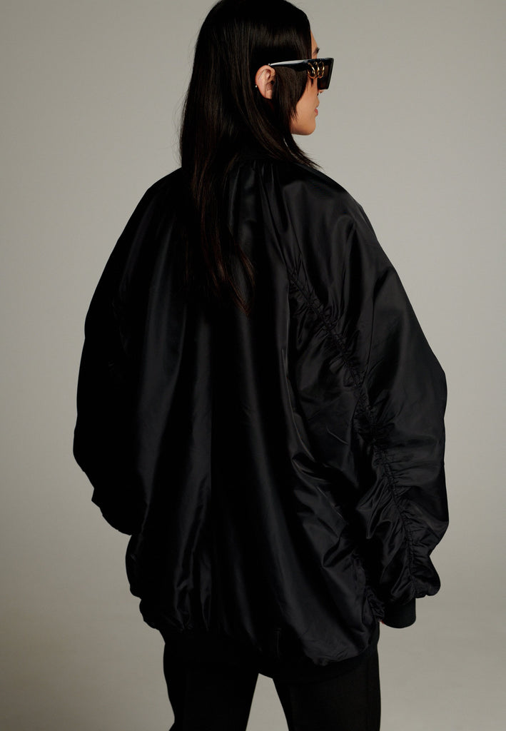 A bomber jacket combines modern utility with sleek style, this one features a unique silver and gold zipper detail, adding a touch of metallic contrast to its minimalist design. The bomber silhouette offers a relaxed, casual vibe, while its durable nylon fabric ensures both comfort and functionality. This versatile jacket effortlessly blends style and practicality, making it a timeless addition to any wardrobe.