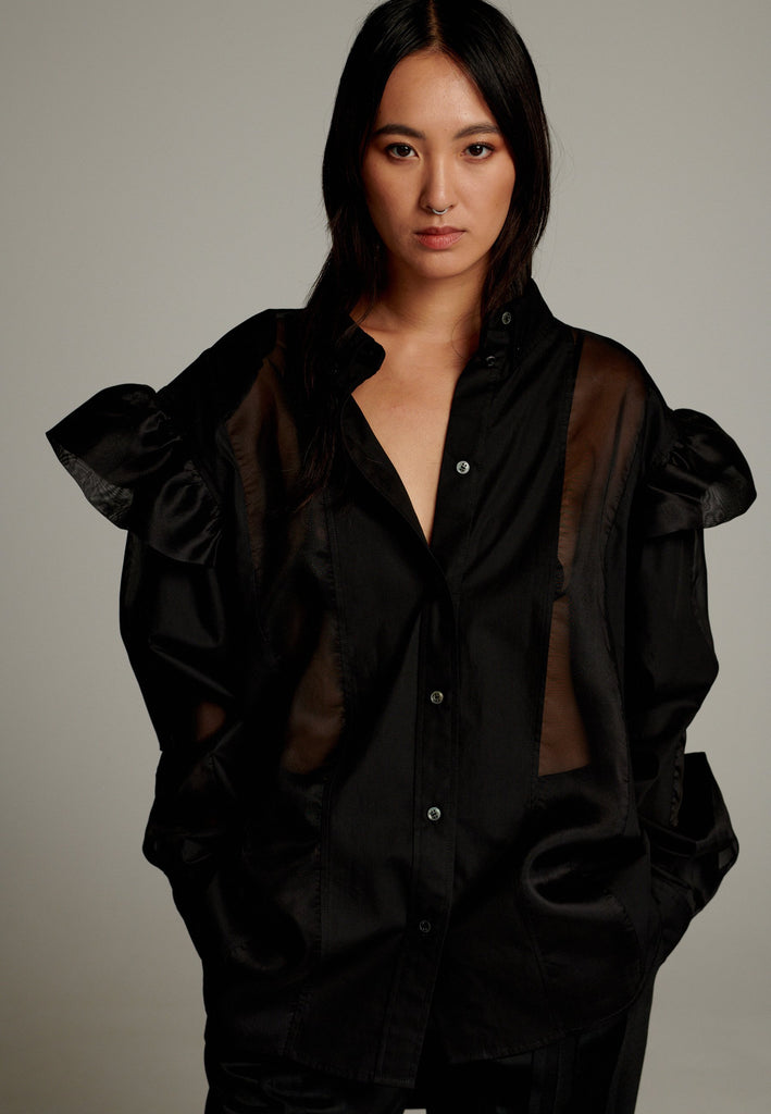The oversized fit offers comfort and versatility, and also combines modern edge with a touch of elegance. Designed with sheer transparent panels, it adds a subtle, stylish contrast to the solid fabric. Ruffled details on the shoulders enhance its playful, feminine appeal while maintaining a relaxed, contemporary vibe.