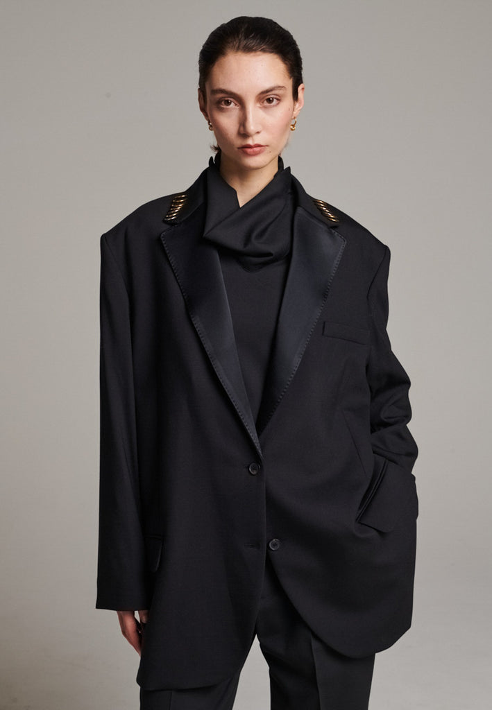A classic yet oversized blazer, designed to make a bold statement while maintaining timeless appeal. The lower portion of the lapels is crafted from a contrasting shiny fabric, adding an unexpected touch of texture and luxury. The upper part of the collar area is beautifully embellished with gold and silver rings at the tips, offering a refined, modern twist. This piece combines traditional tailoring with avant-garde details, perfect for elevating any outfit with sophistication and edge.