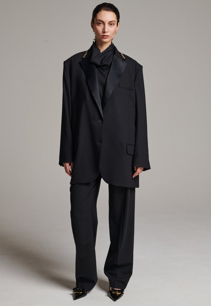 A classic yet oversized blazer, designed to make a bold statement while maintaining timeless appeal. The lower portion of the lapels is crafted from a contrasting shiny fabric, adding an unexpected touch of texture and luxury. The upper part of the collar area is beautifully embellished with gold and silver rings at the tips, offering a refined, modern twist. This piece combines traditional tailoring with avant-garde details, perfect for elevating any outfit with sophistication and edge.