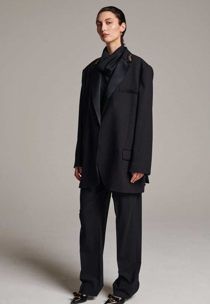 A classic yet oversized blazer, designed to make a bold statement while maintaining timeless appeal. The lower portion of the lapels is crafted from a contrasting shiny fabric, adding an unexpected touch of texture and luxury. The upper part of the collar area is beautifully embellished with gold and silver rings at the tips, offering a refined, modern twist. This piece combines traditional tailoring with avant-garde details, perfect for elevating any outfit with sophistication and edge.