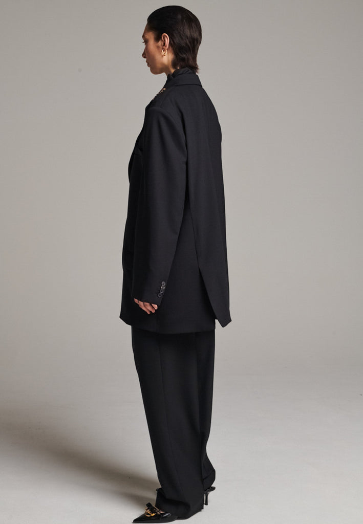 A classic yet oversized blazer, designed to make a bold statement while maintaining timeless appeal. The lower portion of the lapels is crafted from a contrasting shiny fabric, adding an unexpected touch of texture and luxury. The upper part of the collar area is beautifully embellished with gold and silver rings at the tips, offering a refined, modern twist. This piece combines traditional tailoring with avant-garde details, perfect for elevating any outfit with sophistication and edge.