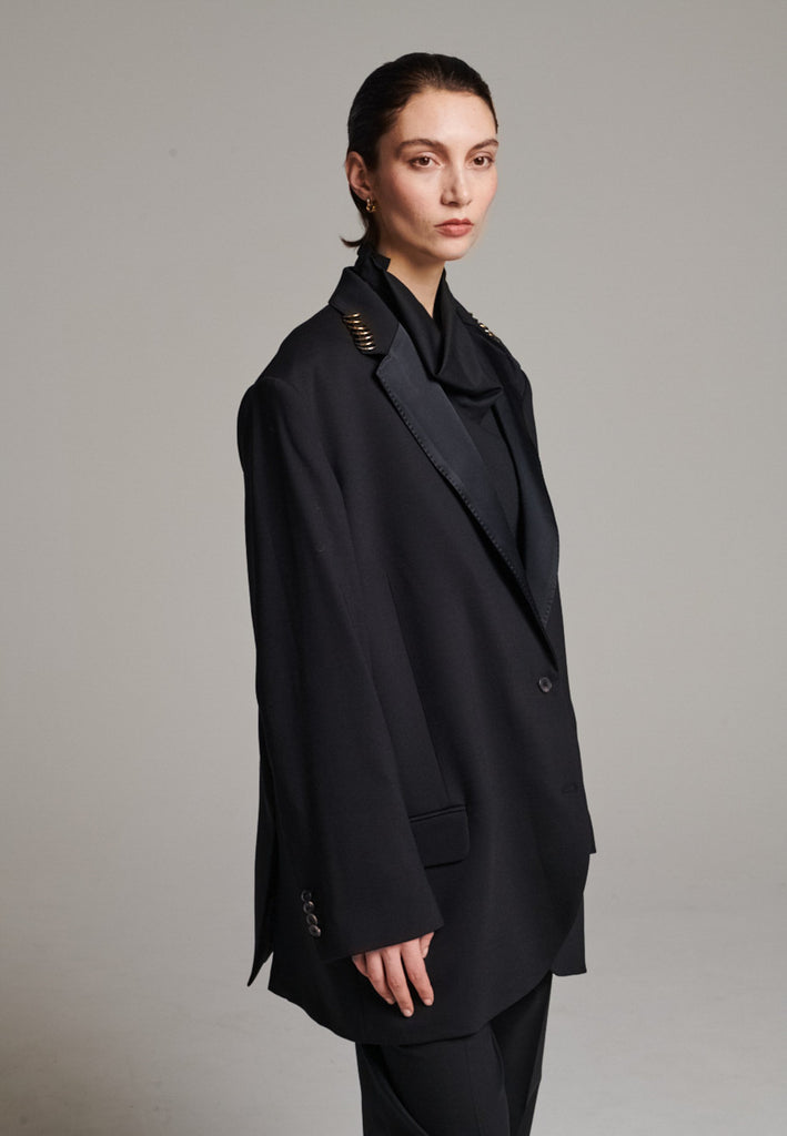 A classic yet oversized blazer, designed to make a bold statement while maintaining timeless appeal. The lower portion of the lapels is crafted from a contrasting shiny fabric, adding an unexpected touch of texture and luxury. The upper part of the collar area is beautifully embellished with gold and silver rings at the tips, offering a refined, modern twist. This piece combines traditional tailoring with avant-garde details, perfect for elevating any outfit with sophistication and edge.