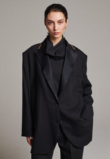 A classic yet oversized blazer, designed to make a bold statement while maintaining timeless appeal. The lower portion of the lapels is crafted from a contrasting shiny fabric, adding an unexpected touch of texture and luxury. The upper part of the collar area is beautifully embellished with gold and silver rings at the tips, offering a refined, modern twist. This piece combines traditional tailoring with avant-garde details, perfect for elevating any outfit with sophistication and edge.