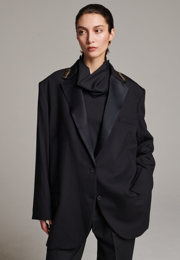 A classic yet oversized blazer, designed to make a bold statement while maintaining timeless appeal. The lower portion of the lapels is crafted from a contrasting shiny fabric, adding an unexpected touch of texture and luxury. The upper part of the collar area is beautifully embellished with gold and silver rings at the tips, offering a refined, modern twist. This piece combines traditional tailoring with avant-garde details, perfect for elevating any outfit with sophistication and edge.