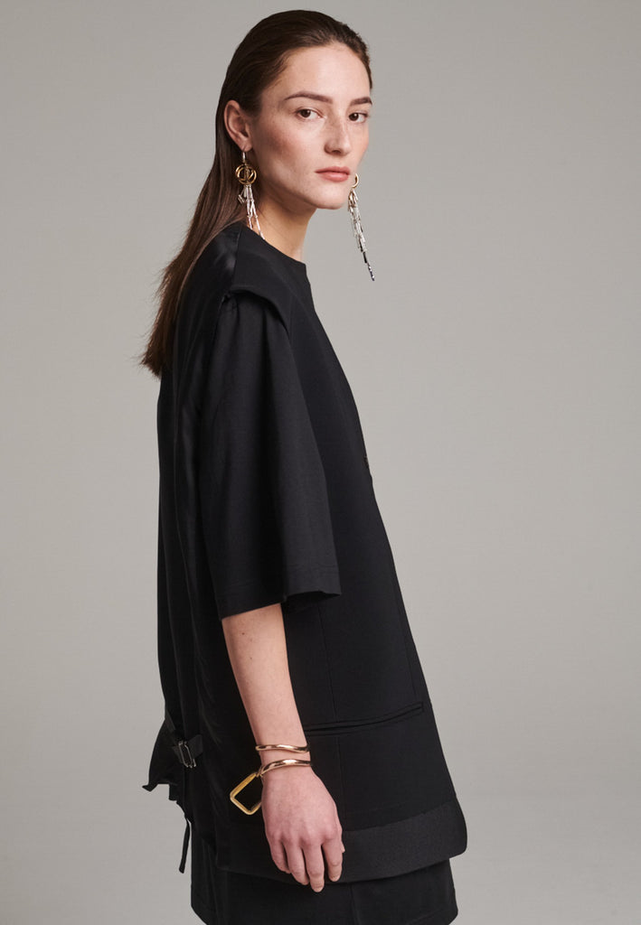 A bold reinterpretation of a classic vest silhouette, cut in a voluminous, oversized shape, it combines sharp tailoring with a contemporary twist. A standout feature is the exaggerated hem, crafted in a contrasting black fabric with a subtle sheen, adding depth and visual interest to the minimalist design. With its clean lines, deep armholes, and striking finish, this piece layers effortlessly and brings a refined edge to both casual and dressed-up looks.