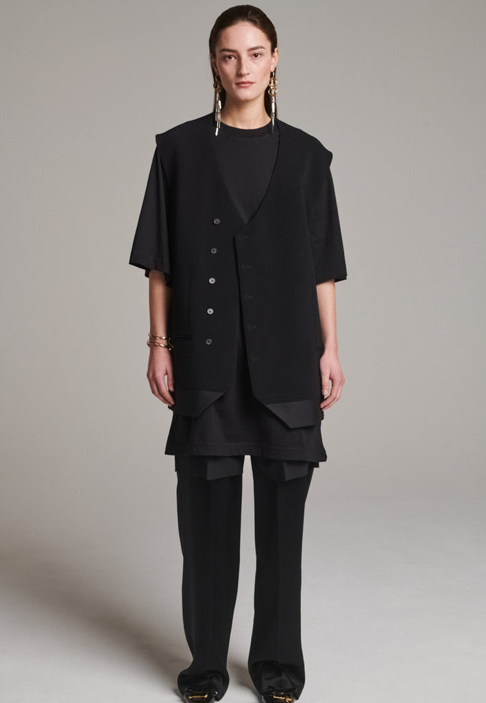 A bold reinterpretation of a classic vest silhouette, cut in a voluminous, oversized shape, it combines sharp tailoring with a contemporary twist. A standout feature is the exaggerated hem, crafted in a contrasting black fabric with a subtle sheen, adding depth and visual interest to the minimalist design. With its clean lines, deep armholes, and striking finish, this piece layers effortlessly and brings a refined edge to both casual and dressed-up looks.