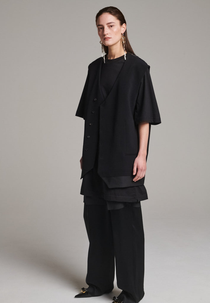 A bold reinterpretation of a classic vest silhouette, cut in a voluminous, oversized shape, it combines sharp tailoring with a contemporary twist. A standout feature is the exaggerated hem, crafted in a contrasting black fabric with a subtle sheen, adding depth and visual interest to the minimalist design. With its clean lines, deep armholes, and striking finish, this piece layers effortlessly and brings a refined edge to both casual and dressed-up looks.