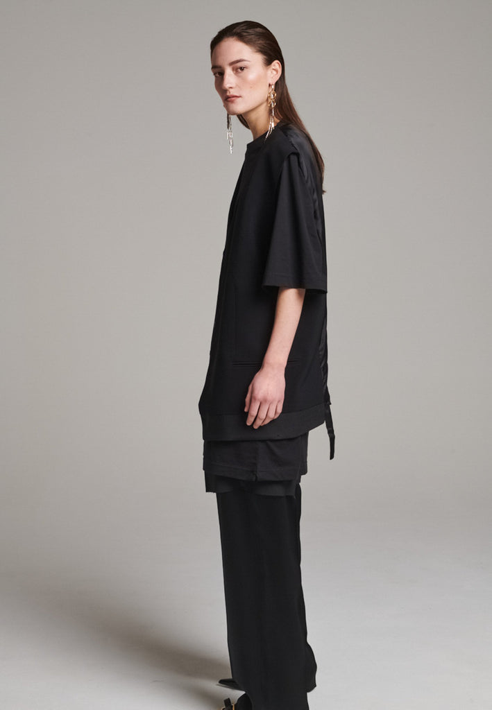A bold reinterpretation of a classic vest silhouette, cut in a voluminous, oversized shape, it combines sharp tailoring with a contemporary twist. A standout feature is the exaggerated hem, crafted in a contrasting black fabric with a subtle sheen, adding depth and visual interest to the minimalist design. With its clean lines, deep armholes, and striking finish, this piece layers effortlessly and brings a refined edge to both casual and dressed-up looks.
