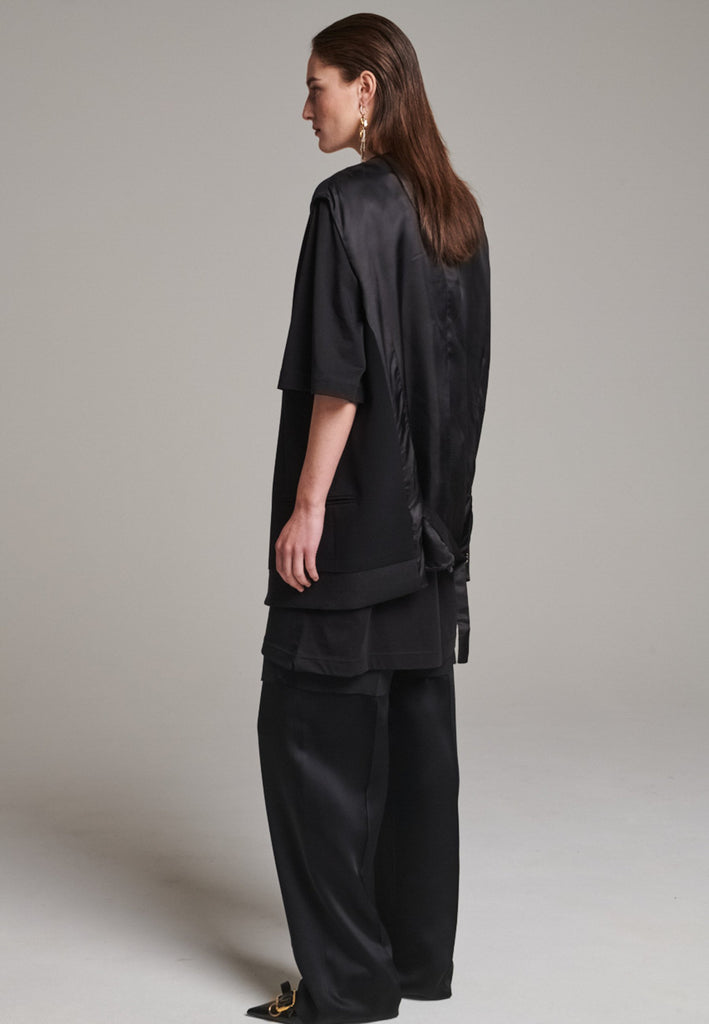 A bold reinterpretation of a classic vest silhouette, cut in a voluminous, oversized shape, it combines sharp tailoring with a contemporary twist. A standout feature is the exaggerated hem, crafted in a contrasting black fabric with a subtle sheen, adding depth and visual interest to the minimalist design. With its clean lines, deep armholes, and striking finish, this piece layers effortlessly and brings a refined edge to both casual and dressed-up looks.