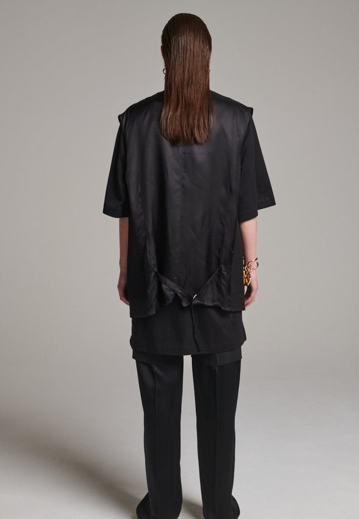 A bold reinterpretation of a classic vest silhouette, cut in a voluminous, oversized shape, it combines sharp tailoring with a contemporary twist. A standout feature is the exaggerated hem, crafted in a contrasting black fabric with a subtle sheen, adding depth and visual interest to the minimalist design. With its clean lines, deep armholes, and striking finish, this piece layers effortlessly and brings a refined edge to both casual and dressed-up looks.