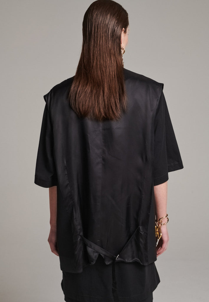 A bold reinterpretation of a classic vest silhouette, cut in a voluminous, oversized shape, it combines sharp tailoring with a contemporary twist. A standout feature is the exaggerated hem, crafted in a contrasting black fabric with a subtle sheen, adding depth and visual interest to the minimalist design. With its clean lines, deep armholes, and striking finish, this piece layers effortlessly and brings a refined edge to both casual and dressed-up looks.
