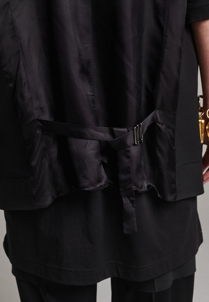 A bold reinterpretation of a classic vest silhouette, cut in a voluminous, oversized shape, it combines sharp tailoring with a contemporary twist. A standout feature is the exaggerated hem, crafted in a contrasting black fabric with a subtle sheen, adding depth and visual interest to the minimalist design. With its clean lines, deep armholes, and striking finish, this piece layers effortlessly and brings a refined edge to both casual and dressed-up looks.