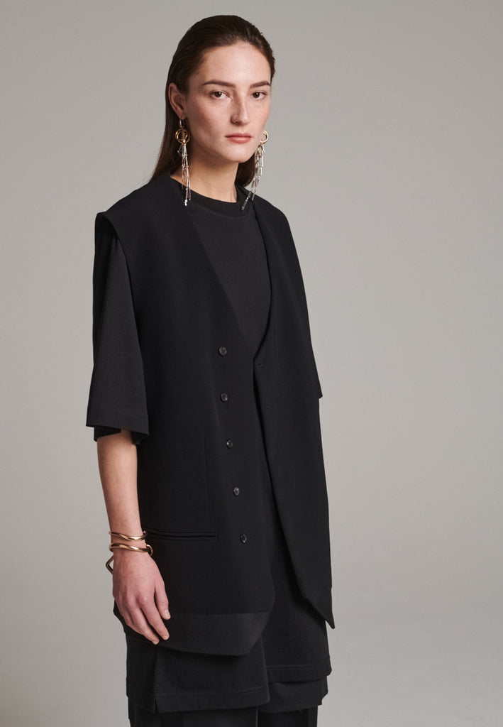 A bold reinterpretation of a classic vest silhouette, cut in a voluminous, oversized shape, it combines sharp tailoring with a contemporary twist. A standout feature is the exaggerated hem, crafted in a contrasting black fabric with a subtle sheen, adding depth and visual interest to the minimalist design. With its clean lines, deep armholes, and striking finish, this piece layers effortlessly and brings a refined edge to both casual and dressed-up looks.