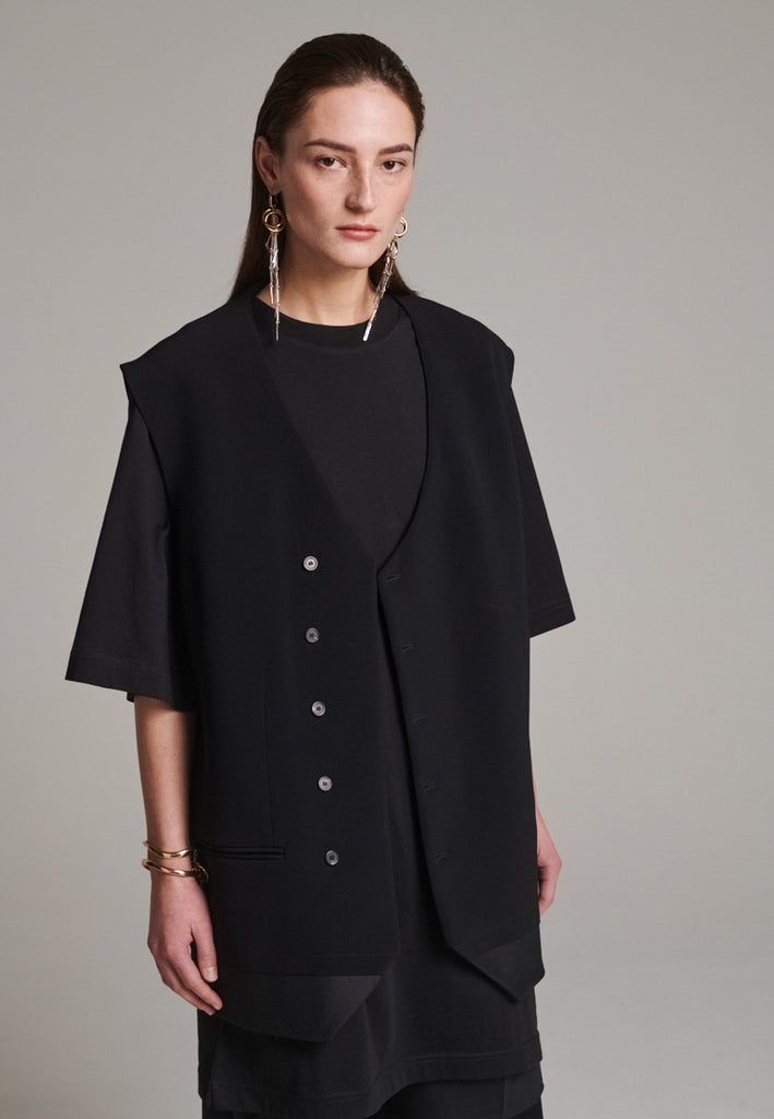 A bold reinterpretation of a classic vest silhouette, cut in a voluminous, oversized shape, it combines sharp tailoring with a contemporary twist. A standout feature is the exaggerated hem, crafted in a contrasting black fabric with a subtle sheen, adding depth and visual interest to the minimalist design. With its clean lines, deep armholes, and striking finish, this piece layers effortlessly and brings a refined edge to both casual and dressed-up looks.