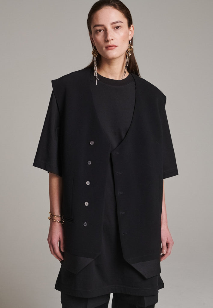A bold reinterpretation of a classic vest silhouette, cut in a voluminous, oversized shape, it combines sharp tailoring with a contemporary twist. A standout feature is the exaggerated hem, crafted in a contrasting black fabric with a subtle sheen, adding depth and visual interest to the minimalist design. With its clean lines, deep armholes, and striking finish, this piece layers effortlessly and brings a refined edge to both casual and dressed-up looks.