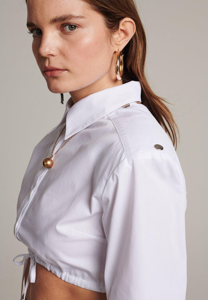 FRENKEN's Best shirt is a directional take on a classic. Cut from white crisp cotton-poplin, it's cropped and gathered at the bust. It has a thin cord closure at the bottom. Sharply padded shoulders detailed with push buttons.