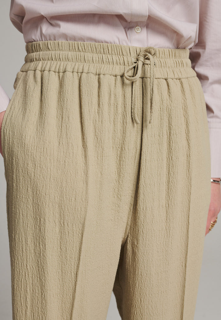Cropped flowy pants in stone with a high-rise. Designed with a loose relaxed fit and drawstring flexible waistband. Pressed front pleat to create a more sophisticated look. Detailed with a welt pocket at the back.