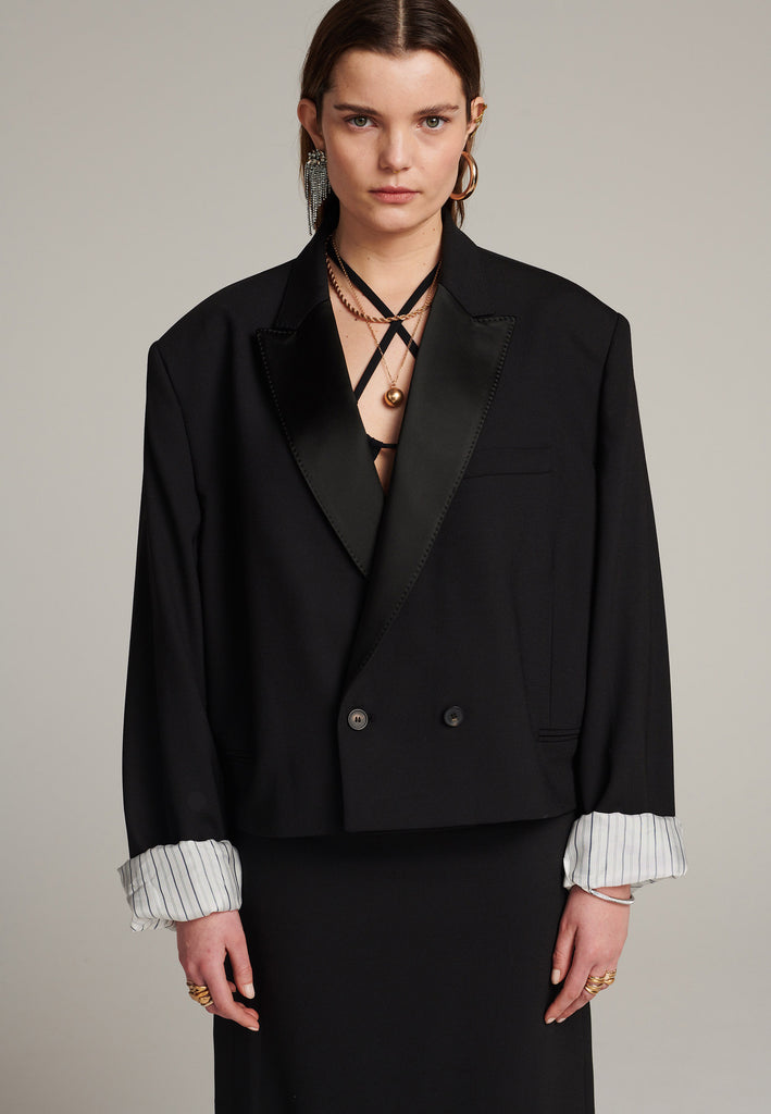 Menswear-inspired smoking blazer with satin lapel, horn buttons, pockets, and shoulder pads for an extreme oversized fitting. The exaggerated long sleeves can be rolled up to reveal a contrasting striped lining. Cut from a fine wool blend, its cropped shape adds a sophisticated touch.