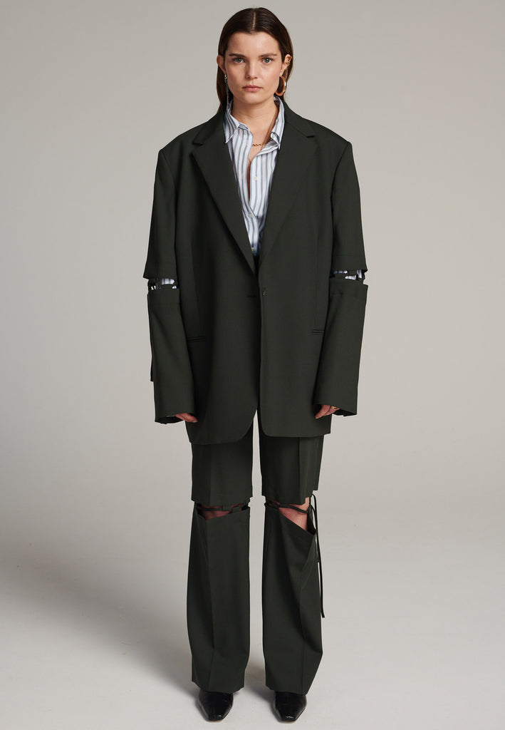 Menswear-inspired smoking blazer in dark green with horn buttons and shoulder pads, emphasizing the oversized fitting. The exaggerated long sleeves can be rolled up to reveal a striped lining. The blazer features a cut-out section around the elbow, making a two-piece sleeve held together by a finely laced string. Can be worn elegantly open, loose or tied together.