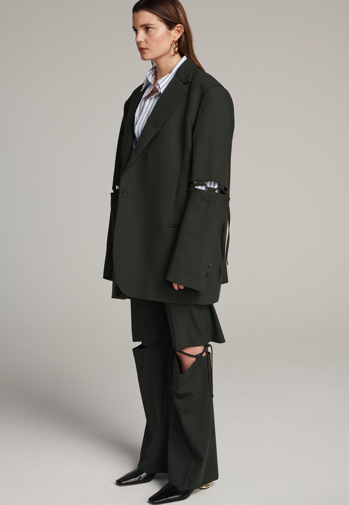 Menswear-inspired smoking blazer in dark green with horn buttons and shoulder pads, emphasizing the oversized fitting. The exaggerated long sleeves can be rolled up to reveal a striped lining. The blazer features a cut-out section around the elbow, making a two-piece sleeve held together by a finely laced string. Can be worn elegantly open, loose or tied together.