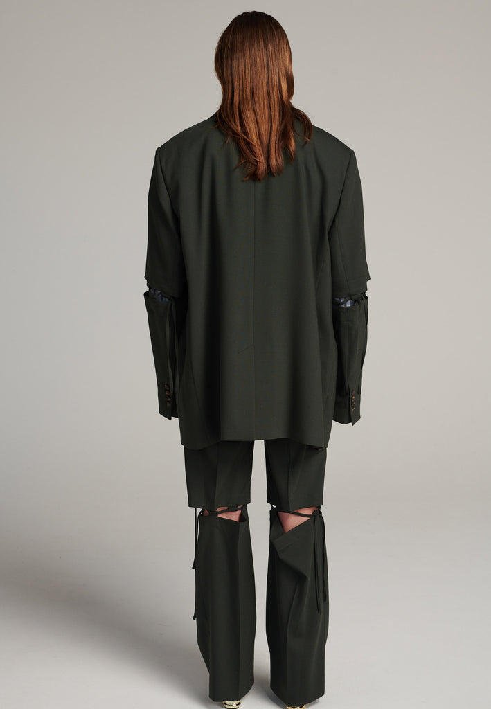 Menswear-inspired smoking blazer in dark green with horn buttons and shoulder pads, emphasizing the oversized fitting. The exaggerated long sleeves can be rolled up to reveal a striped lining. The blazer features a cut-out section around the elbow, making a two-piece sleeve held together by a finely laced string. Can be worn elegantly open, loose or tied together.
