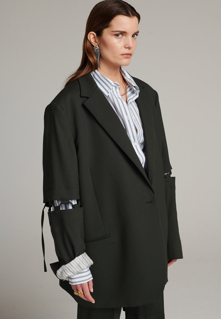 Menswear-inspired smoking blazer in dark green with horn buttons and shoulder pads, emphasizing the oversized fitting. The exaggerated long sleeves can be rolled up to reveal a striped lining. The blazer features a cut-out section around the elbow, making a two-piece sleeve held together by a finely laced string. Can be worn elegantly open, loose or tied together.
