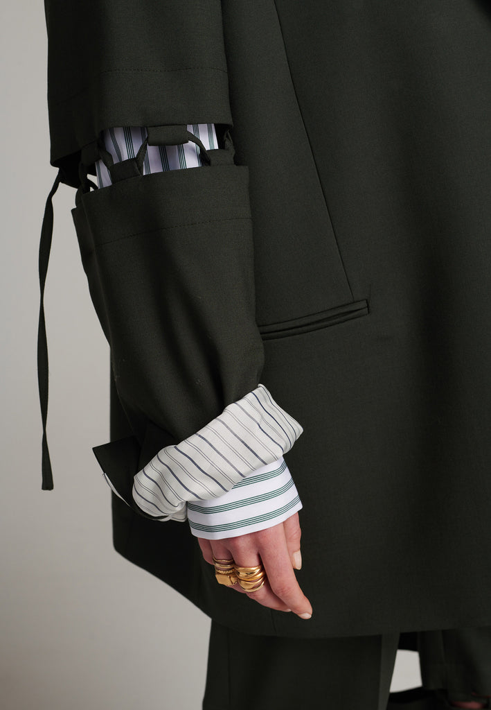 Menswear-inspired smoking blazer in dark green with horn buttons and shoulder pads, emphasizing the oversized fitting. The exaggerated long sleeves can be rolled up to reveal a striped lining. The blazer features a cut-out section around the elbow, making a two-piece sleeve held together by a finely laced string. Can be worn elegantly open, loose or tied together.