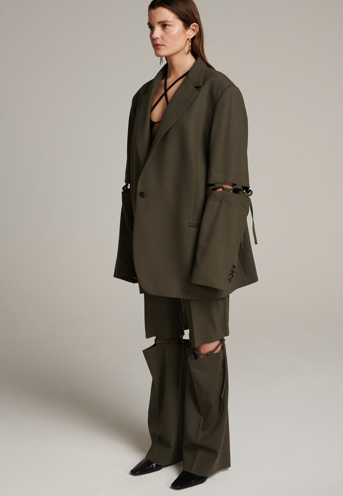 Menswear-inspired smoking blazer in dark camel with horn buttons and shoulder pads, emphasizing the oversized fitting. The exaggerated long sleeves can be rolled up to reveal a striped lining. The blazer features a cut-out section around the elbow, making a two-piece sleeve held together by a finely laced string. Can be worn elegantly open, loose or tied together.