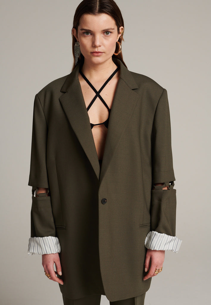 Menswear-inspired smoking blazer in dark camel with horn buttons and shoulder pads, emphasizing the oversized fitting. The exaggerated long sleeves can be rolled up to reveal a striped lining. The blazer features a cut-out section around the elbow, making a two-piece sleeve held together by a finely laced string. Can be worn elegantly open, loose or tied together.