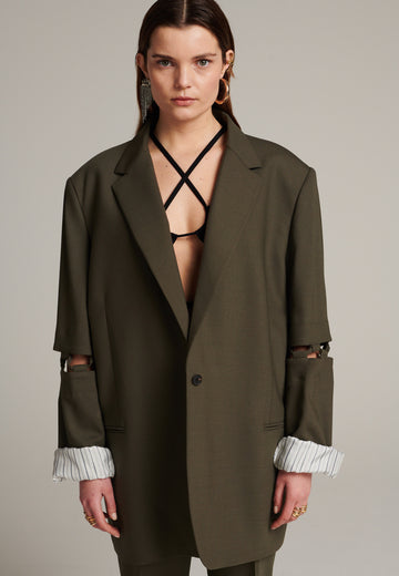Menswear-inspired smoking blazer in dark camel with horn buttons and shoulder pads, emphasizing the oversized fitting. The exaggerated long sleeves can be rolled up to reveal a striped lining. The blazer features a cut-out section around the elbow, making a two-piece sleeve held together by a finely laced string. Can be worn elegantly open, loose or tied together.