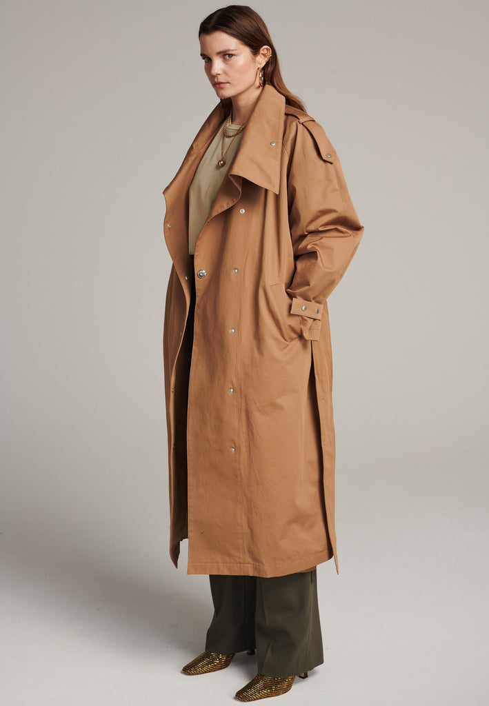 Oversized trench coat tailored from crisp camel cotton for a loose shape. Detailed with silver metal press buttons at front closure at the cuffs. An oversized collar to hide in and a self-fabric belt to cinch the loose oversized fit.