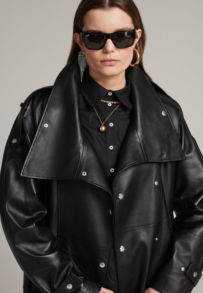 Oversized trench coat crafted in butterly soft lamb leather in black. Detailed with silver metal press buttons at front closure, epaulettes at shoulders and cuffs. Designed with a maxi collar with the snap-fastening buttons and a self-leather belt tp cinch the loose oversized fit.