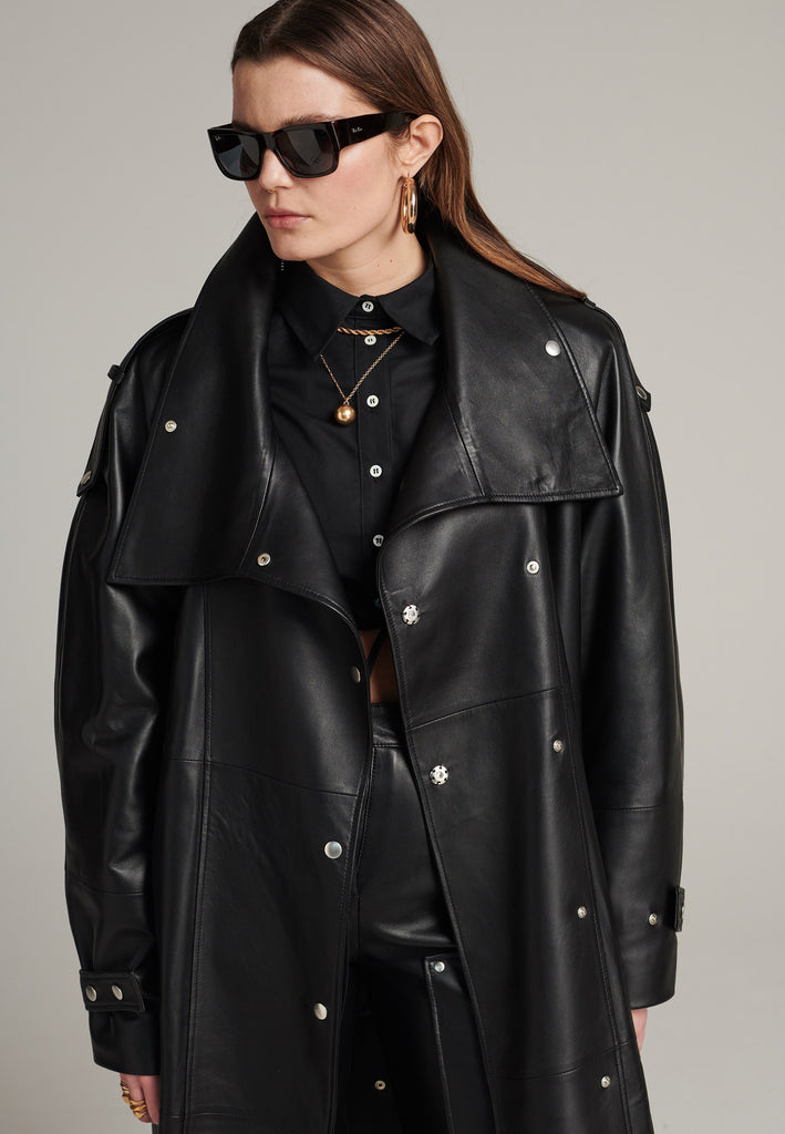 Oversized trench coat crafted in butterly soft lamb leather in black. Detailed with silver metal press buttons at front closure, epaulettes at shoulders and cuffs. Designed with a maxi collar with the snap-fastening buttons and a self-leather belt tp cinch the loose oversized fit.