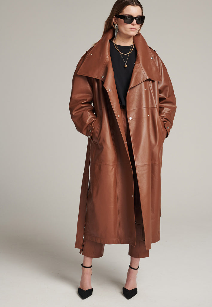 Oversized trench coat crafted in butterly soft lamb leather in cognac. Detailed with silver metal press buttons at front closure, epaulettes at shoulders and cuffs. Designed with a maxi collar with the snap-fastening buttons and a self-leather belt tp cinch the loose oversized fit.