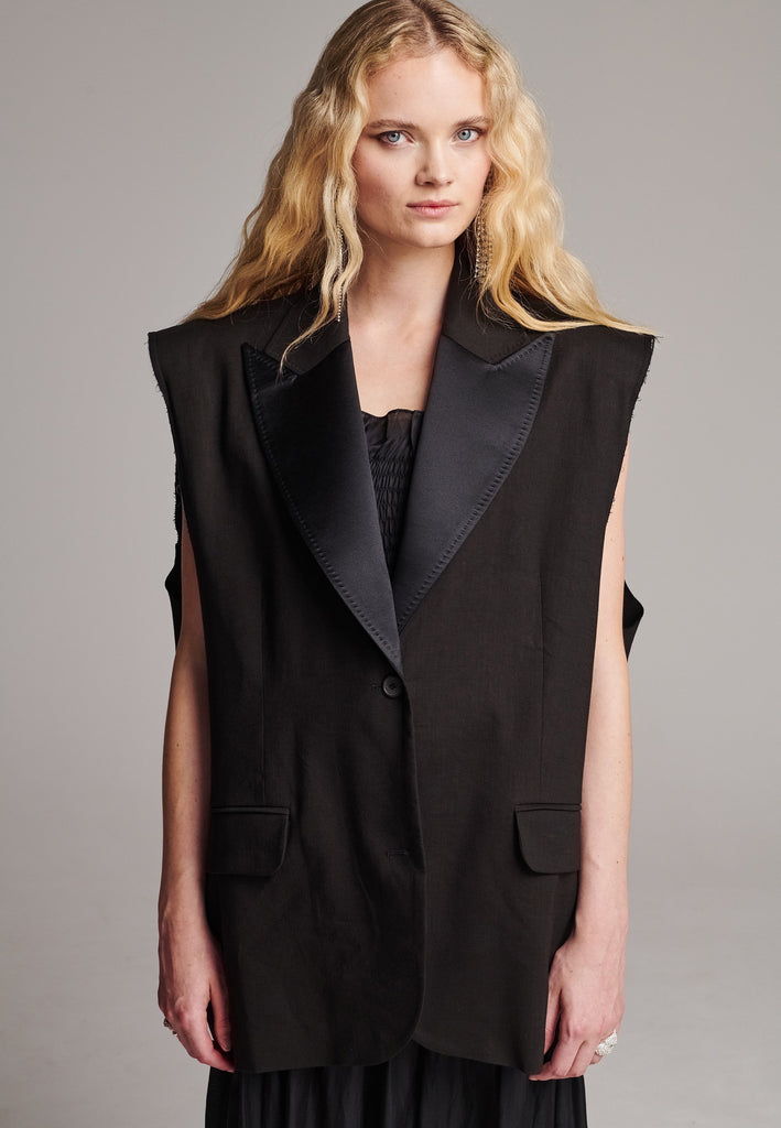 Oversized sleeveless blazer. Padded shoulders, horn button closure, detailed with darts and flap pockets. Raw edge armhole that elevates the piece’s statement. Central back vent, inside welt pocket, tailored with tuxedo satin lapels finished by and needle stitching. Cut from soft summer wool blend and lined with viscose satin.