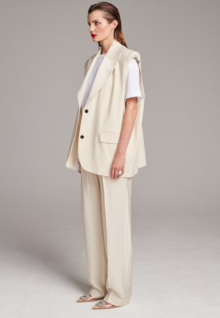 Oversized sleeveless blazer. Padded shoulders, horn button closure, detailed with darts and flap pockets. Raw edge armhole that elevates the piece’s statement. Central back vent, inside welt pocket, tailored with tuxedo satin lapels finished by needle stitching. Cut from a soft summer wool blend and lined with viscose satin.
