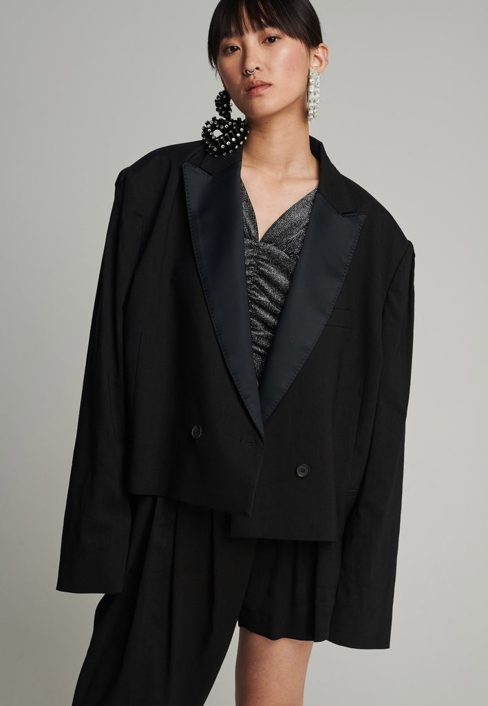 Wide-shouldered short blazer in black. Features shoulder-pads, pockets, and exaggerated long sleeves. Fits oversize