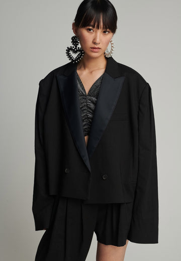 Wide-shouldered short blazer in black. Features shoulder-pads, pockets, and exaggerated long sleeves. Fits oversize