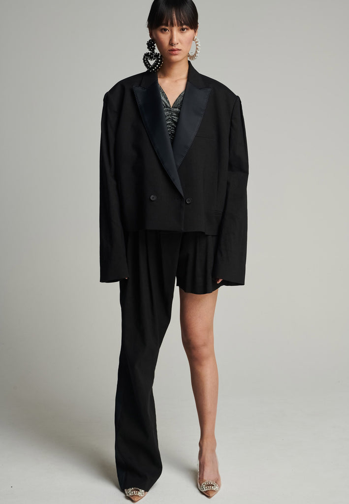 Wide-shouldered short blazer in black. Features shoulder-pads, pockets, and exaggerated long sleeves. Fits oversize
