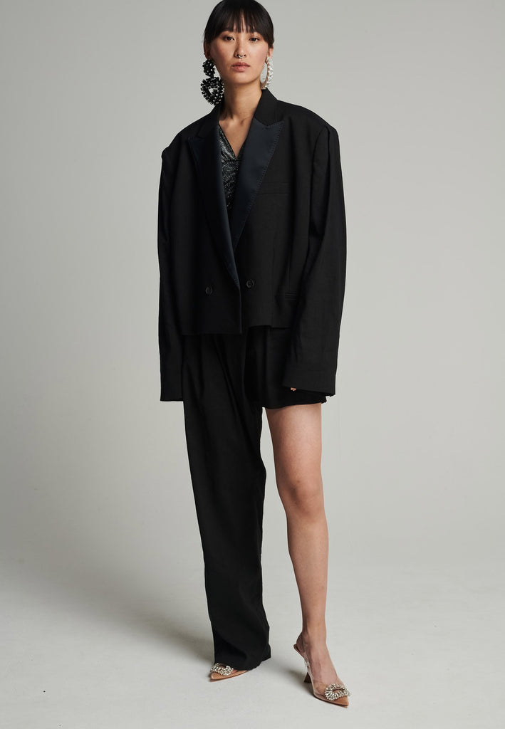 Wide-shouldered short blazer in black. Features shoulder-pads, pockets, and exaggerated long sleeves. Fits oversize