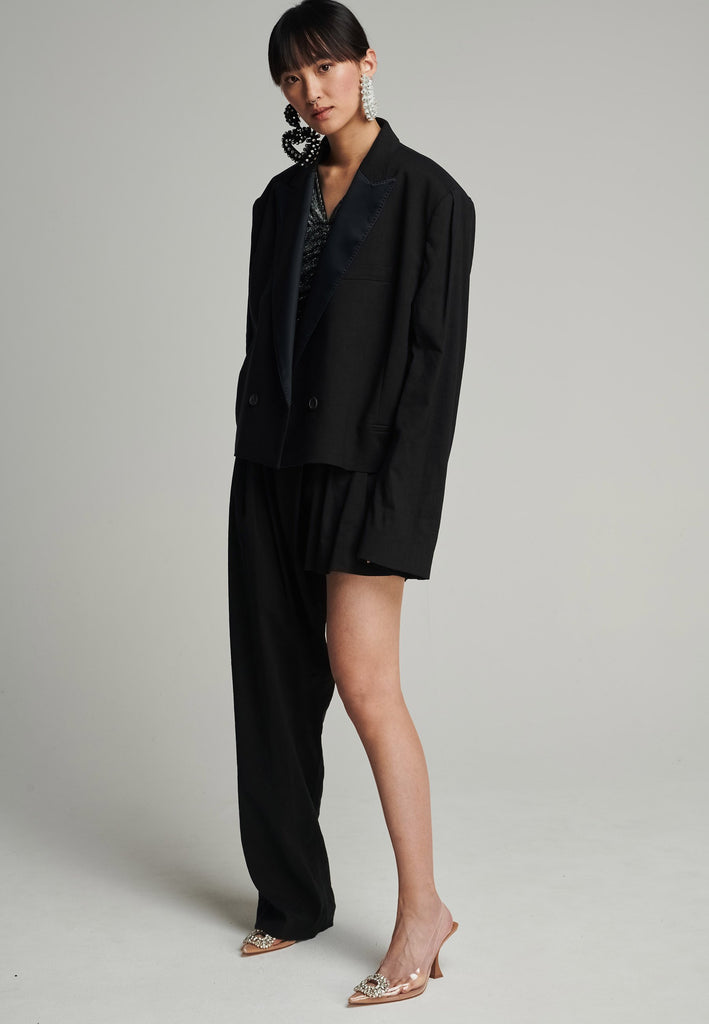 Wide-shouldered short blazer in black. Features shoulder-pads, pockets, and exaggerated long sleeves. Fits oversize